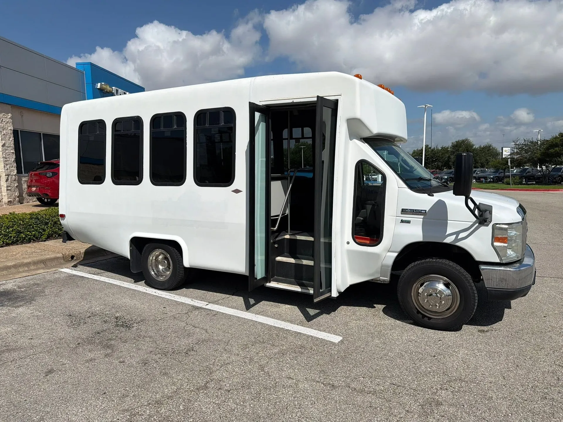 Real 2013 Ford E-350 minibus with the passenger door open in a sunlit commercial lot, with a blue building and blue sky behind it
